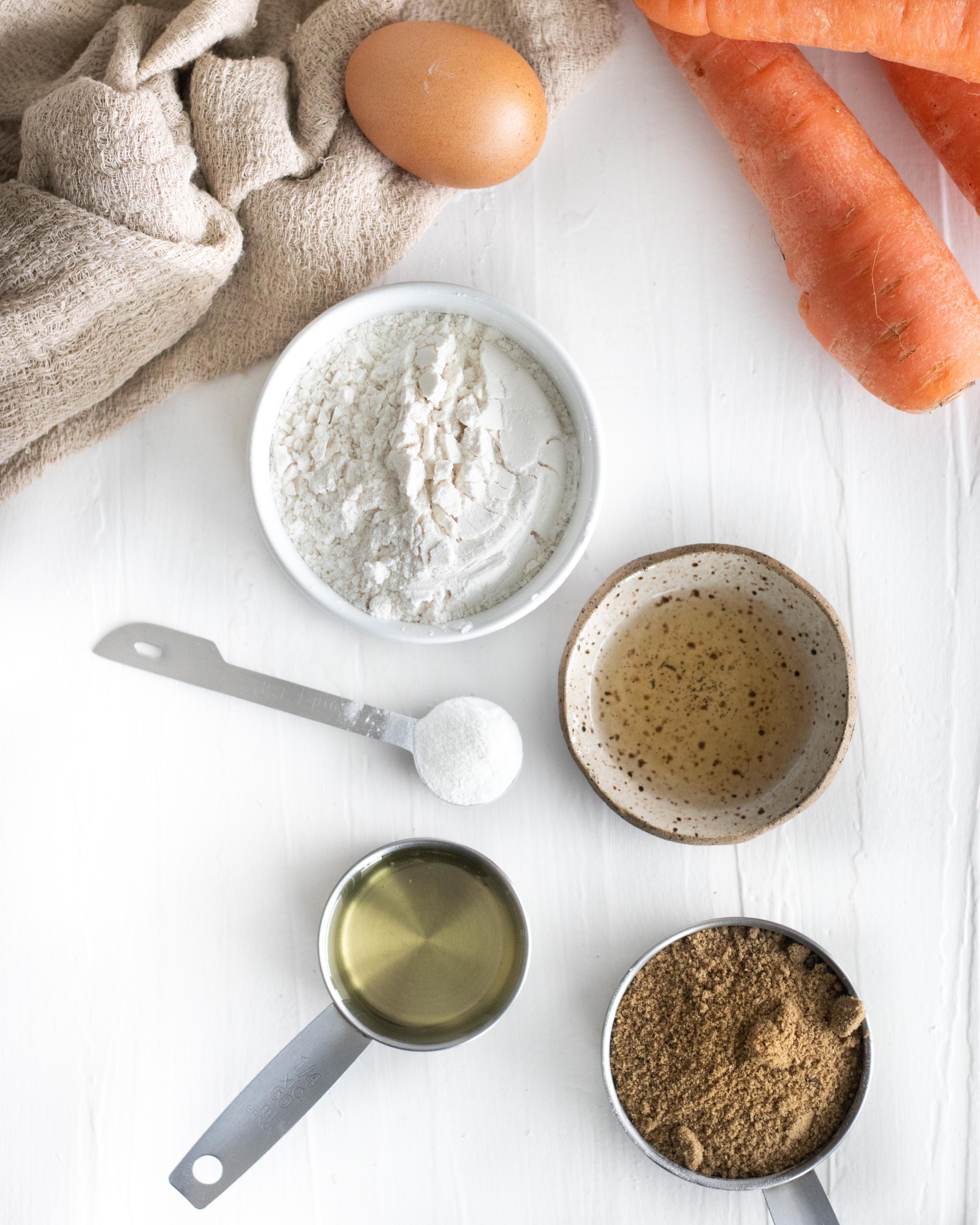 ingredientes dispostos sobre uma mesa branca, contendo cenoura, ovo, óleo, açúcar mascavo, mix de farinha sem glúten, fermento e sal